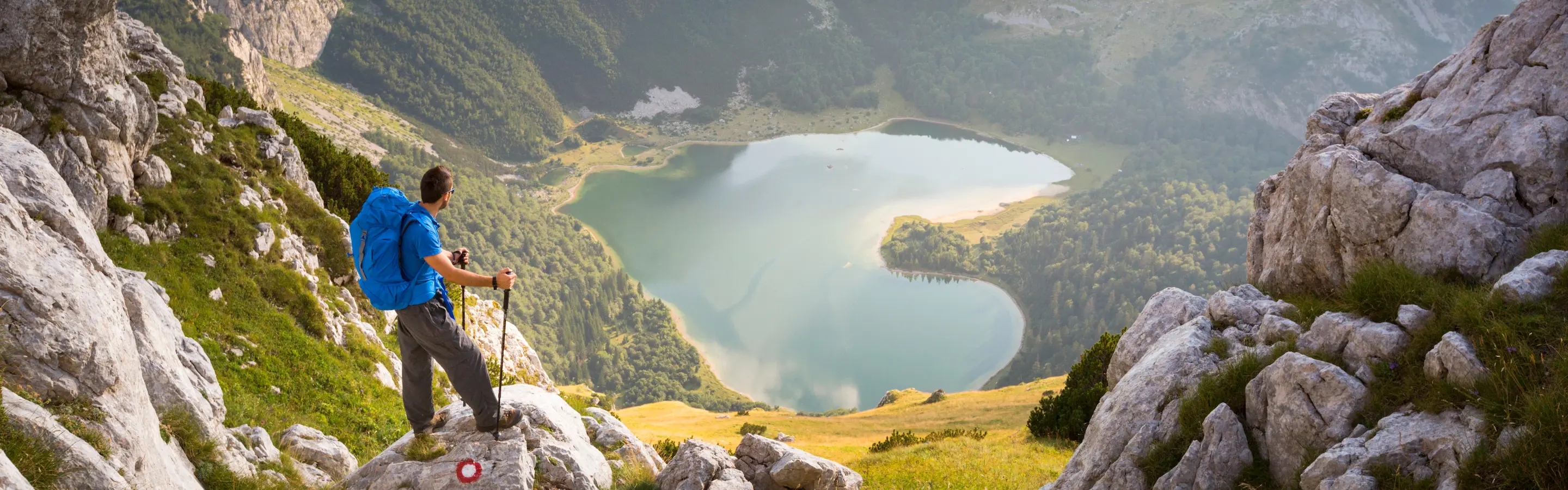 Ein Wanderer mit Rucksack steht auf einem Felsvorsprung und schaut auf den Gletschersee Trnovačko jezero.