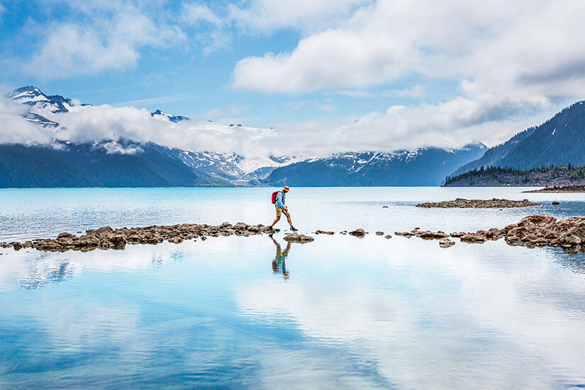 Ein Mann mit Hut und Rucksack springt am Ufer des berühmten Lake Garibaldi von Stein zu Stein über das Wasser. Packliste Kanada Garibaldi