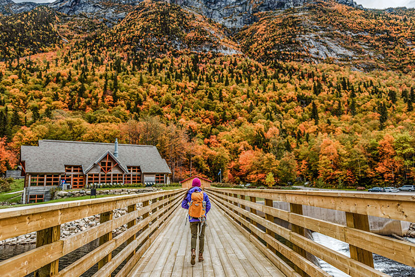 Eine Frau mit Mütze und Rucksack begibt sich in Québec über eine Holzbrücke auf dem Weg zu einem wunderschönen Haus Holz und Felssteinen. Im Hintergrund leuchtet der Wald in lieblichen Tönen aus Gelb, Rot und Orange. Packliste Kanada Quebec