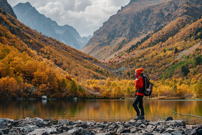 In herbstlicher Szenerie spaziert eine Urlauberin mit Rucksack am steinigen Ufer eines Sees entlang. In unmittelbarer Nähe befindet sich eine Schlucht, die sich aus dem Zusammenschluss dreier majestätischer Berge zusammensetzt. Packliste Kanada Herbst