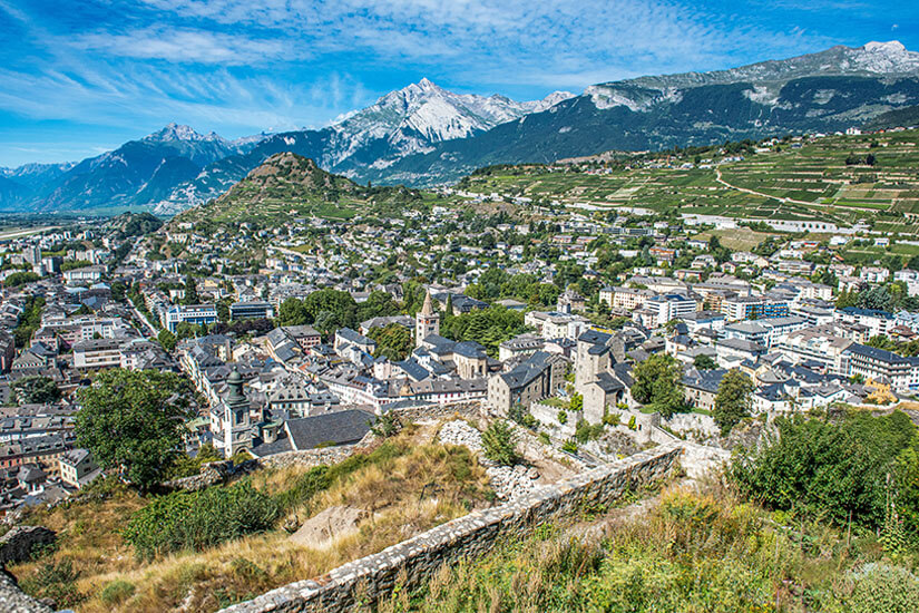 Die Stadt Sion mit verschneitem Berggipfel im Hintergrund. Zwischen den hellen Häusern liegt viel grüne Natur. Sion Schweiz Willkommen