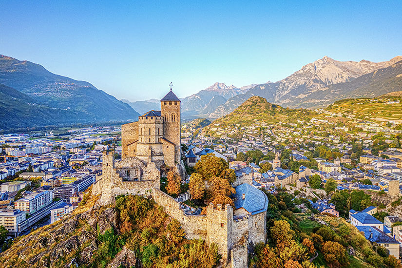 Die Basilika von Sion thront auf einem Hügel, umgeben von herbstlichen Bäumen. Die Stadt und Berggipfel liegen im Hintergrund. Sion Schweiz Basilika