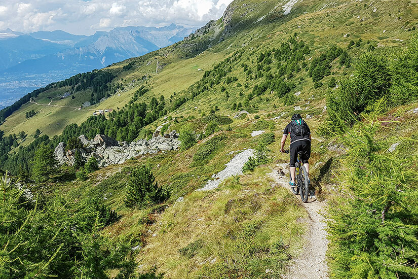 Ein Mountainbiker fährt über einen Pfad in den grünen Bergen vom Wallis. Im fernen Hintergrund ist eine Stadt im Tal zu sehen. Sion Schweiz Mountainbike
