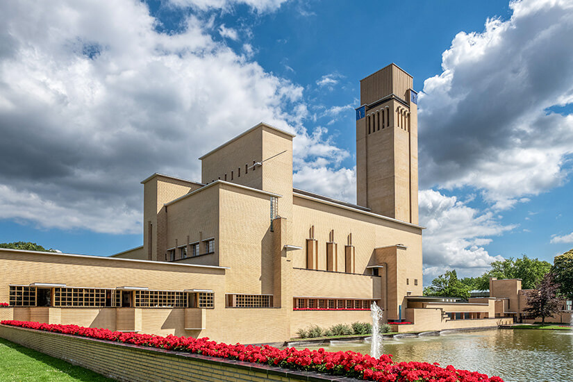 Gebäude aus hellem Stein mit einem hohen Turm, im Vordergrund ist ein Wasserbecken mit Fontäne und rote Blumen zu sehen. Hilversum Rathaus