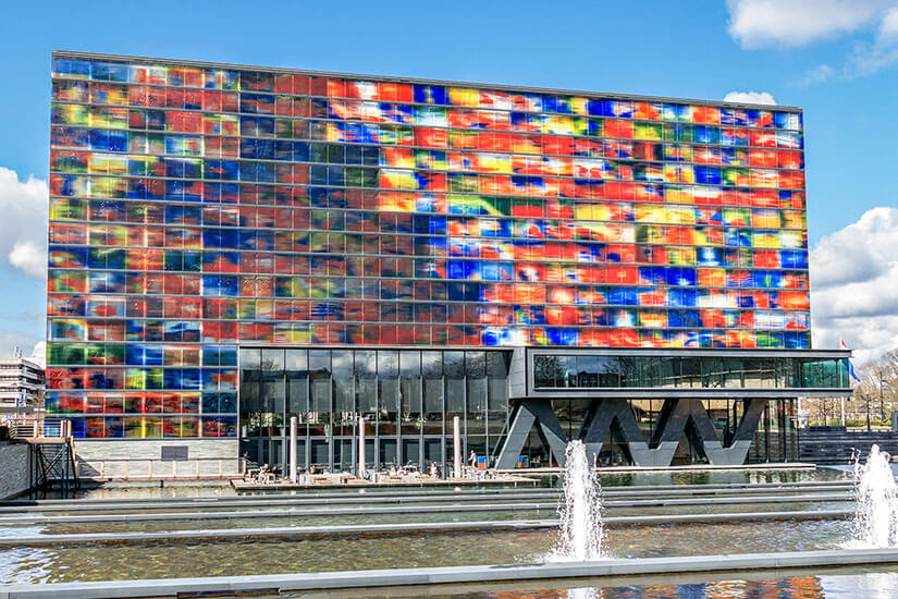 Rechteckiges Gebäude mit bunter Glasfassade, davor ist ein Becken mit Wasserfontäne zu sehen. Hilversum Medienmuseum
