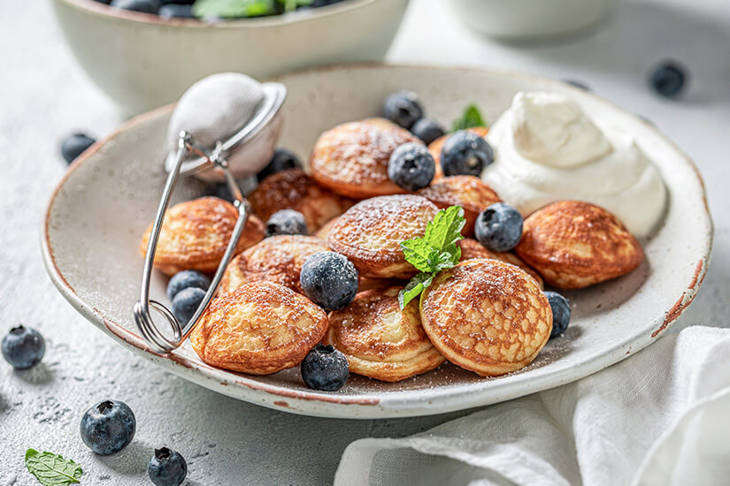 Ein Teller mit kleinen Pfannkuchen, Blaubeeren, Sahne und Puderzucker. Niederlande Poffertjes