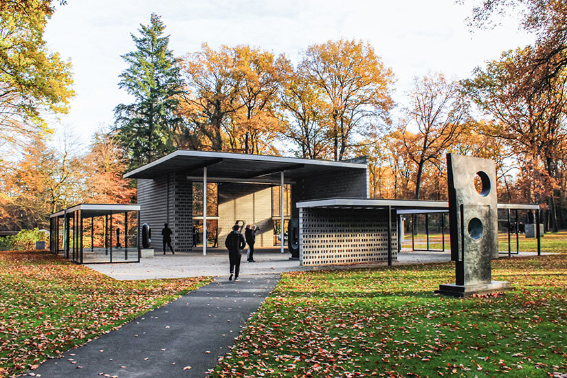 Kleines Gebäude in einem parkähnlichen Gelände mit bunt verfärbten Bäumen im Hintergrund. Davor ist eine Skulptur zu sehen. Hilversum Kunstmuseum