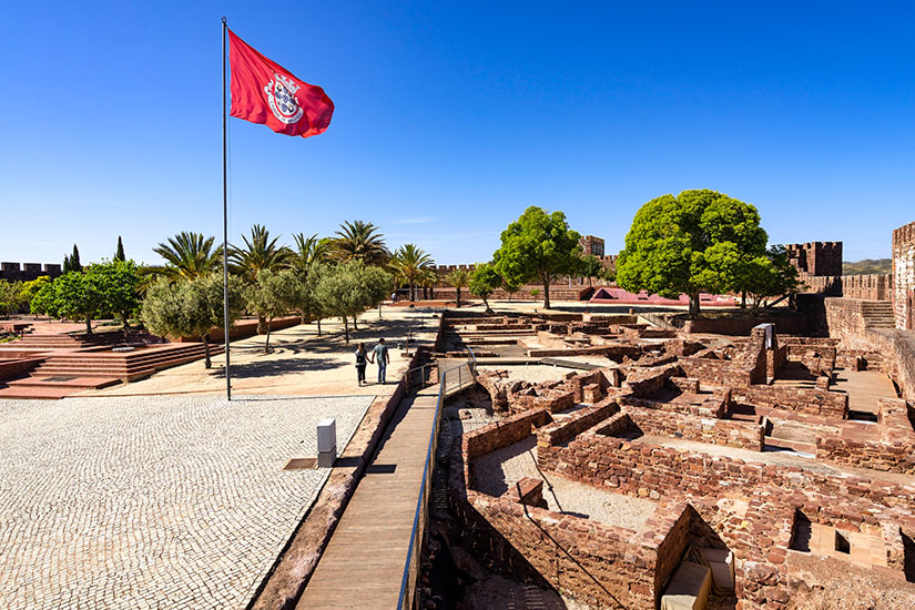 Die roten Mauern einer Burg, links ist eine wehende, rote Fahne zu sehen. Im Hintergrund stehen grüne Bäume und Palmen. Silves Castelo