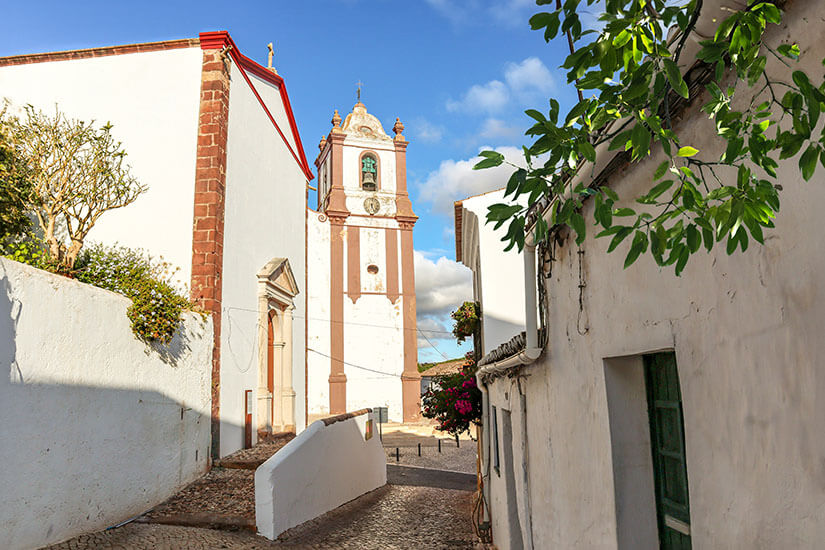 Ein kleine, kopfsteingepflasterte Gasse zwischen weißen Häusern. Im Hintergrund ist der Turm der Kathedrale zu sehen. Silves Gassen