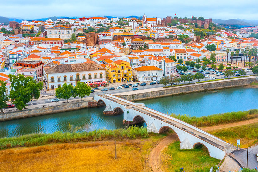 Blick von oben auf eine weiße Brücke mit fünf Bögen, die über einen Fluss führt. Im Hintergrund ist die Stadt zu sehen. Silves Ponte Romana