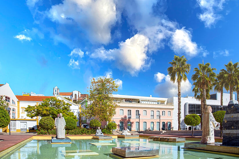 Wasserbecken mit Steinen und Skulpturen. Im Hintergrund sind Palmen und Häuser zu sehen. Silves Park