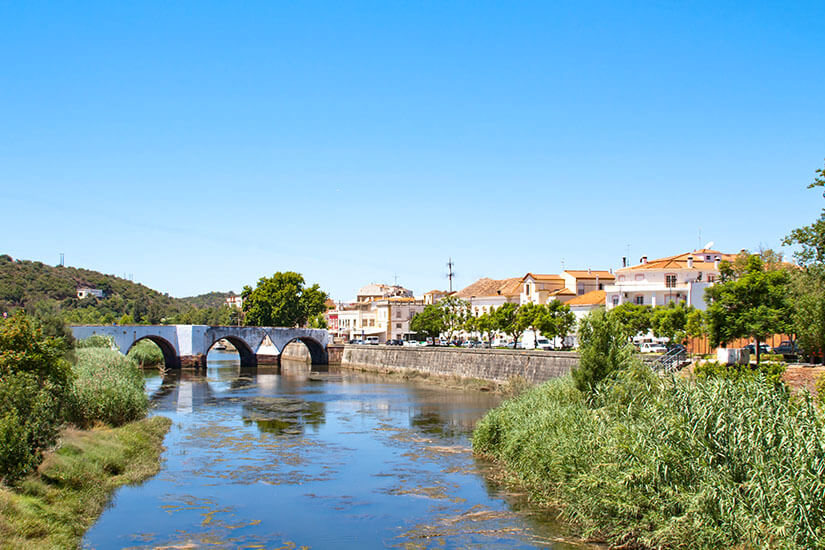 Ein Fluss, der von einer Brücke überquert wird. An den Ufern sind Häuser und grüne Vegetation zu sehen. Portugal Rio Arade