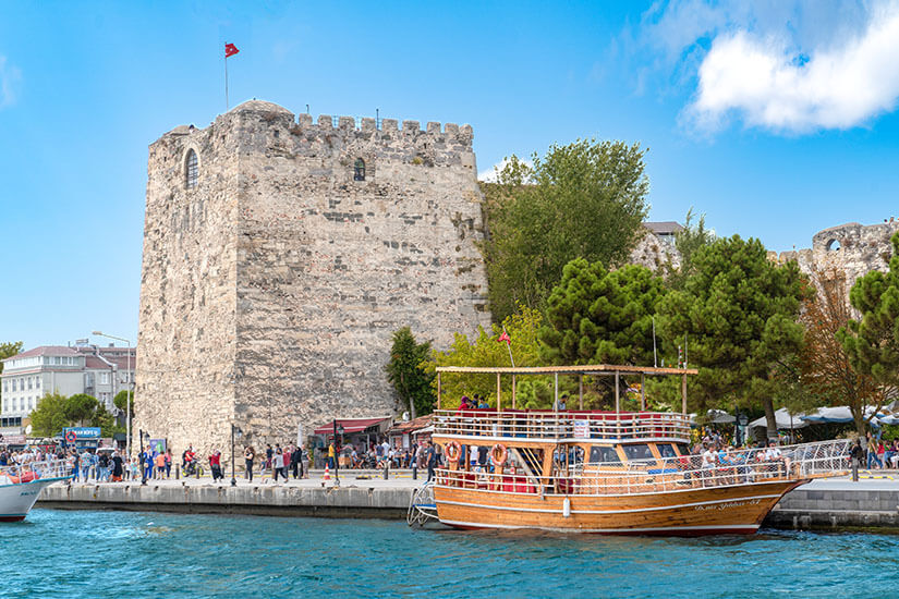 Turm einer Festung an einer Hafenpromenade. Davor ist ein Holzboot zu sehen. Sinop Festung