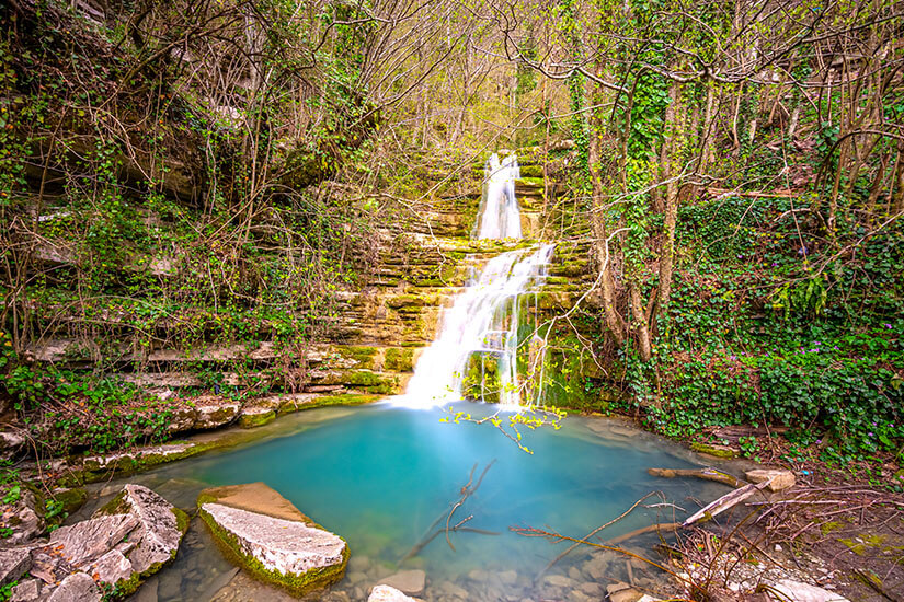 Ein Wasserfall stürzt in ein türkisblaues Becken, an den Seiten sind Bäume zu sehen. Sinop Wasserfall