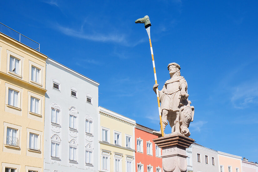 Statue aus Stein auf einem Sockel, dahinter sind bunte Häuserfassaden zu sehen. Ried im Innkreis Hauptplatz