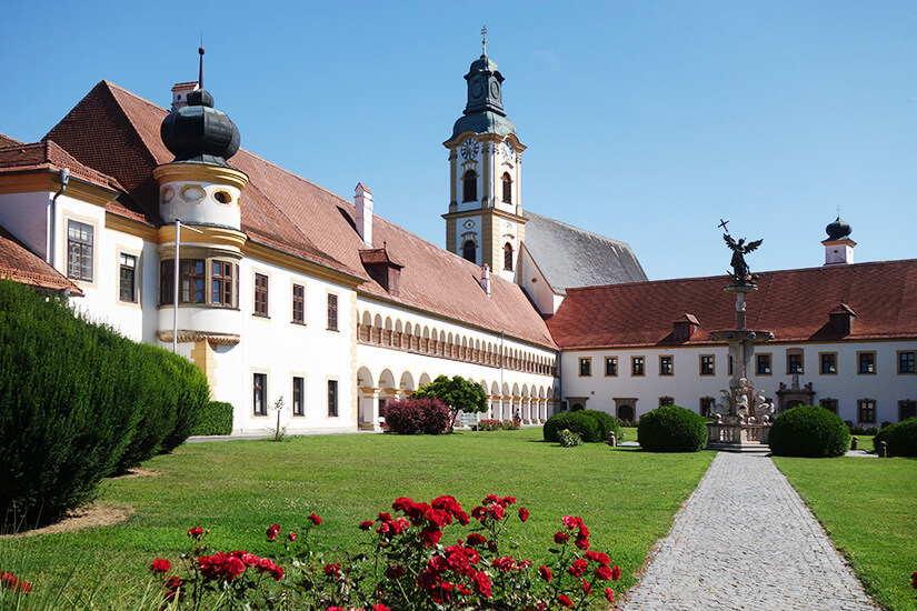 Innenhof eines barocken Klosters mit weißer Fassade mit grünem Rasen. Im Hintergrund ist eine Kirche zu sehen. Ried im Innkreis Kloster