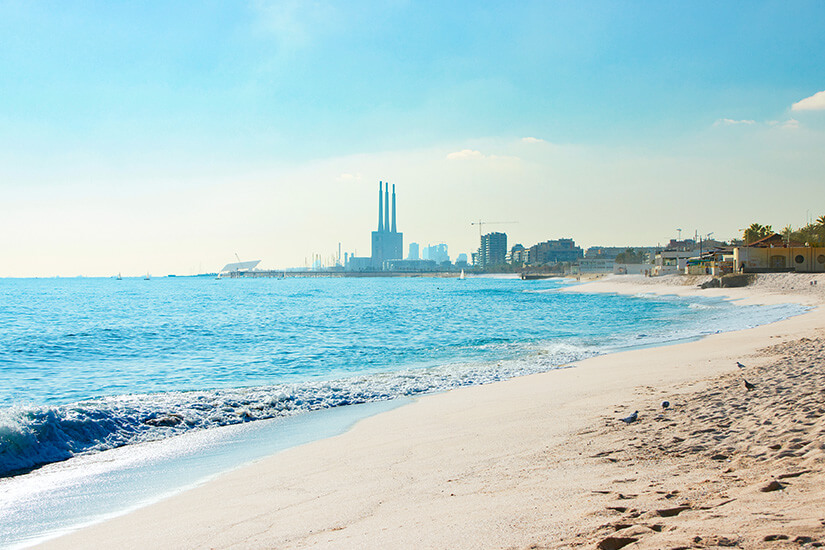 Heller Sandstrand mit kleinen Wellen, im Hintergrund sind drei Türme zu sehen. Badalona Platja del Coco