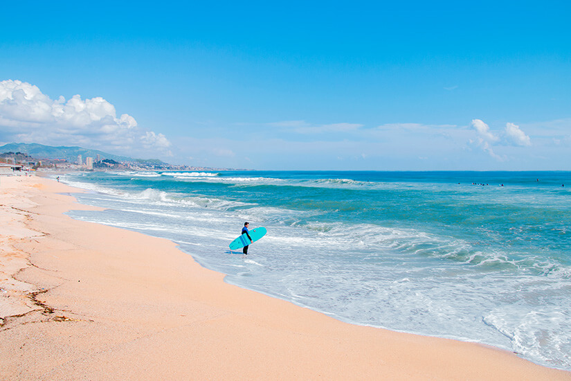 Ein Surfer mit Brett steht an einem Sandstrand. Das türkisblaue Wasser rollt in kleinen Wellen heran. Badalona Surfen