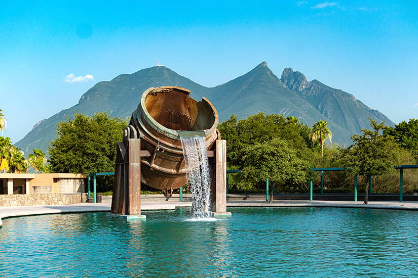 Ein Brunnen, der aus einem riesigen alten Metallkrug geschaffen wurde, gießt Wasser in ein Becken im Fundidora Park. Im Hintergrund liegen Bäume und grüne Berge. Monterrey Mexiko Fundidora