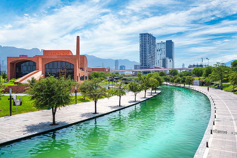 Der idyllische Santa Lucia Riverwalk führt durch einen ruhigen Teil der Stadt. Das Wasser leuchtet türkisblau. Grüne Bäume säumen das Ufer, im Hintergrund sind Wolkenkratzer zu sehen. Monterrey Mexiko Riverwalk