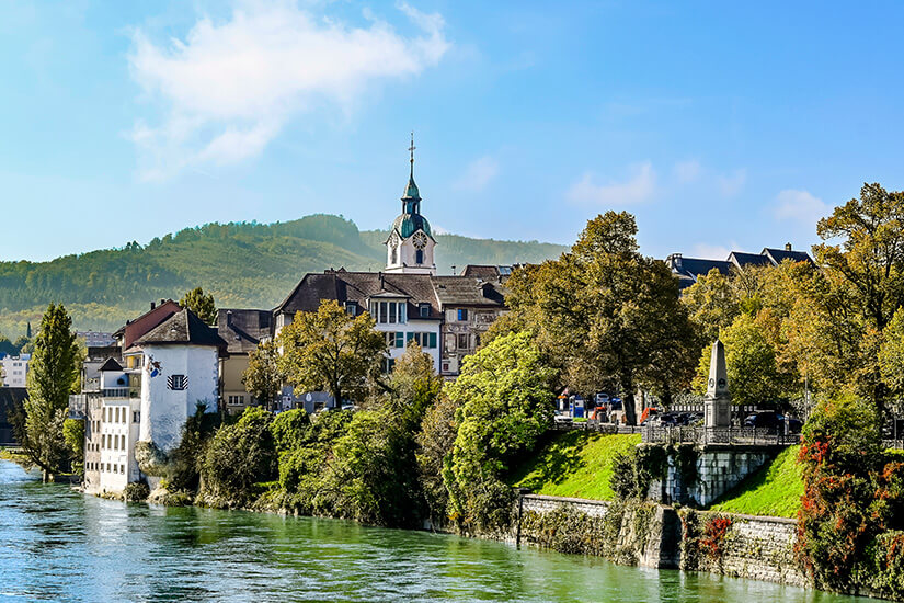 Ein Blick auf Oltens Altstadt vom gegenüberliegenden Flussufer der Aare. Ein Kirchturm und Häuser ragen zwischen grünen Bäumen hervor Olten Schweiz Altstadt