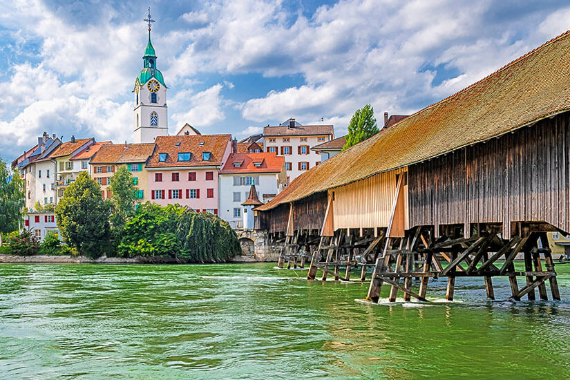Die Alte Holzbrücke erstreckt sich über die Aare in Richtung Altstadt von Olten. Bunte Häuserfassaden und ein Kirchturm sind am anderen Ende zu sehen. Olten Schweiz Bruecke