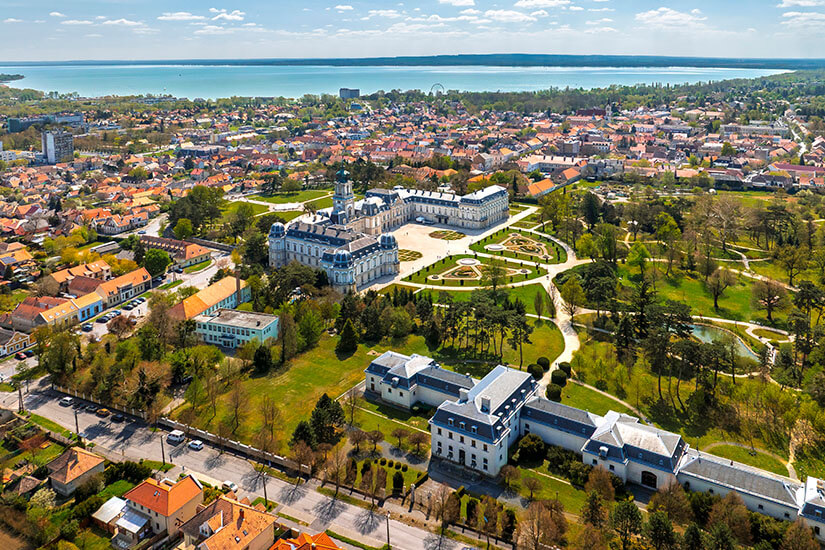 Blick von oben eine Stadt am See, im Vordergrund ist ein weißes Schloss mit grüner Parkanlage zu sehen. Keszthely Stadt