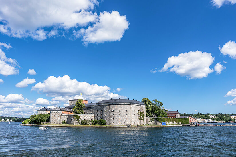 Eine große historische Festungsanlage aus massivem Stein steht auf einer kleinen Insel im Wasser. Darum herum wachsen einige Bäume, und im Hintergrund sind weitere Gebäude sowie Küstenlinien zu erkennen. Vaxholm Festung