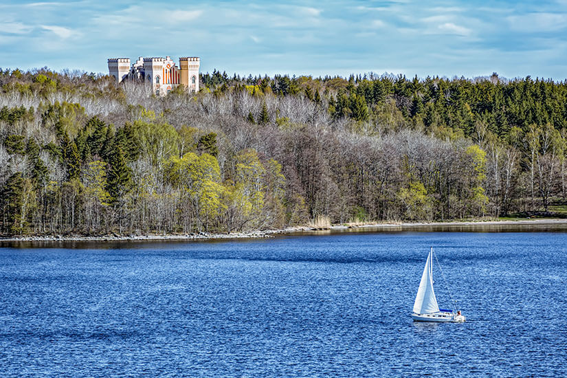 Auf dem Wasser fährt ein kleines Segelboot, das sich vor einer dicht bewaldeten Uferlinie bewegt. Hinter den Bäumen ragt ein großes, helles Schloss mit mehreren Türmen über dem Wald hervor. Vaxholm Bogesund