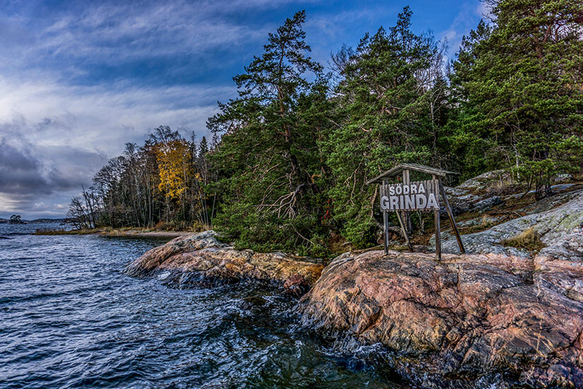 Ein felsiges Ufer mit dunklem, bewegtem Wasser liegt vor einem dichten Wald aus Nadelbäumen. Schweden Grinda Insel