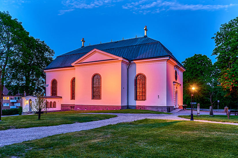 Ein hell erleuchtetes Kirchengebäude mit rosafarbenem Lichtschein steht in einer gepflegten Grünanlage. Der Himmel wirkt nachtblau, und einzelne Laternen sorgen für eine ruhige, stimmungsvolle Atmosphäre. Vaxholm Kirche
