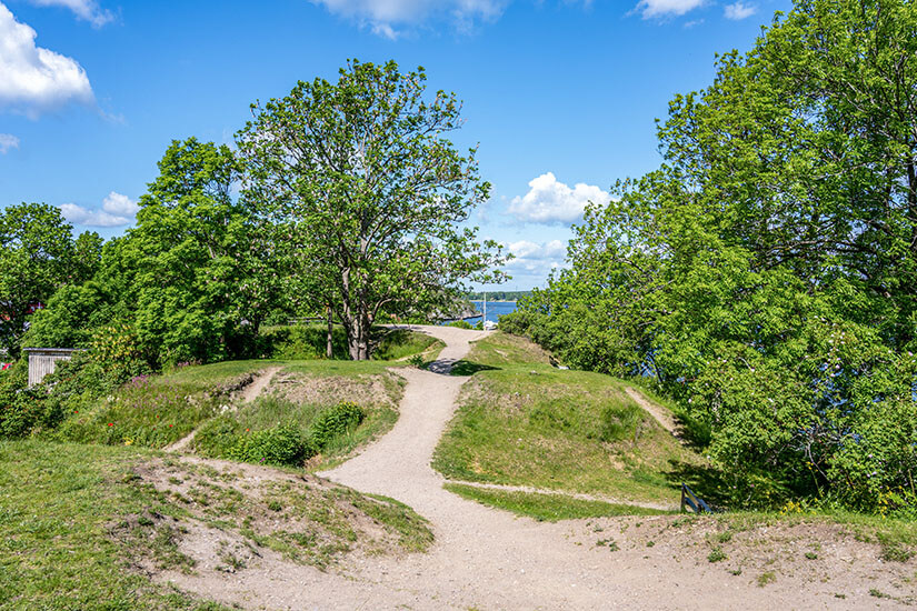 Ein heller Kiesweg führt zwischen grasbewachsenen Hügeln und dichtem, grünen Laub in Richtung Wasser. Mehrere Bäume spenden Schatten und rahmen den Weg angenehm ein. Vaxholm Batteriepark
