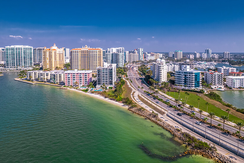 Die Stadt Sarasota mit ihren farbigen Hochhäusern. Eine Straße führt hinein und entlang am grünblauen Meer. Sarasota USA Willkommen