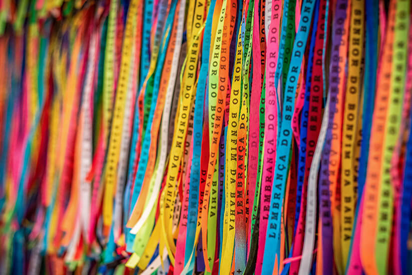Viele bunte, schmale Bänder hängen dicht nebeneinander und bilden ein farbenfrohes Muster. Auf den Stoffstreifen stehen portugiesische Aufschriften, wie sie oft bei religiösen Wunsch- oder Glücksbändern verwendet werden. Souvenirs Brasilien Bonfim