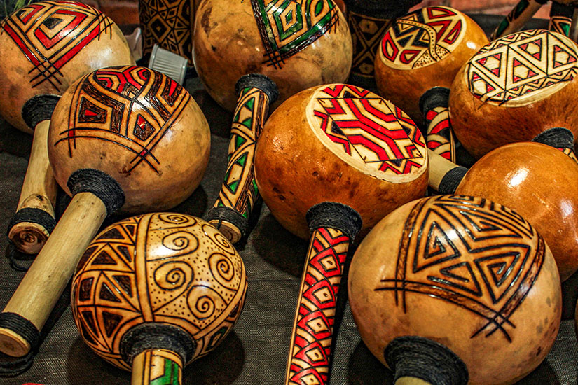 Mehrere verzierte Rasseln liegen eng beieinander, jeweils mit runden, bemalten Köpfen und gemusterten Griffen. Die Ornamente wirken handgearbeitet und zeigen geometrische und ethnisch inspirierte Muster in kräftigen Farben. Souvenirs Brasilien Rumbakugeln