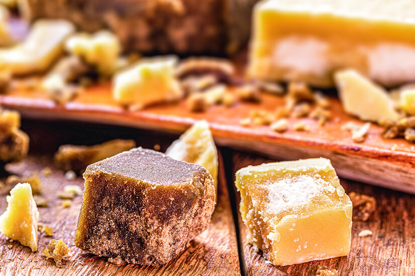 Zwei würfelförmige Stücke aus bräunlicher und heller, krümeliger Masse liegen auf einer hölzernen Oberfläche. Im Hintergrund sind weitere Bruchstücke und größere Blöcke derselben Süßigkeit zu erkennen. Souvenirs Brasilien Rapadura