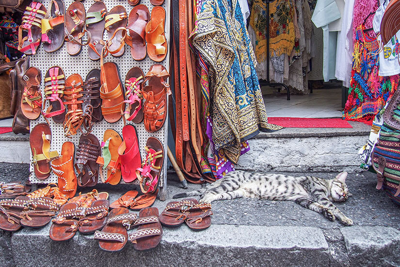 Zahlreiche handgefertigte Ledersandalen sind an einem Verkaufsstand ausgestellt, daneben hängen bunte Stoffe und Gürtel. Vor dem Eingang liegt entspannt eine getigerte Katze auf dem Boden und döst. Sandalen Flip Flops Brasilien