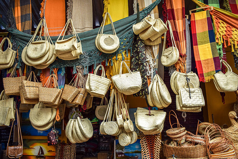 Viele geflochtene Taschen und Körbe aus Naturmaterialien hängen dicht an dicht in verschiedenen Formen und Größen. Darüber sind bunte Tücher und Hängematten dekorativ drapiert und verleihen dem Stand ein lebhaftes Aussehen. Souvenirs Brasilien Basttaschen