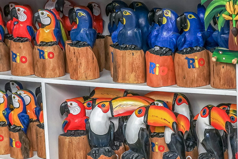 Mehrere bunte, hölzerne Figuren tropischer Vögel – vor allem Papageien und Tukane – stehen dicht nebeneinander in Regalen. Souvenirs Brasilien Holzfiguren