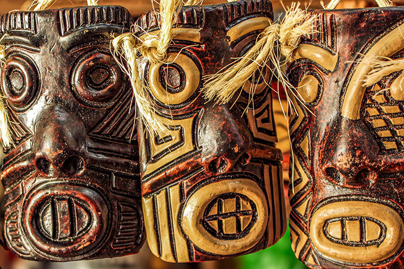 Drei kunstvoll gestaltete Masken hängen nebeneinander und zeigen markante, stilisierte Gesichter mit großen Augen und betonten Mündern. Masken Ton Brasilien