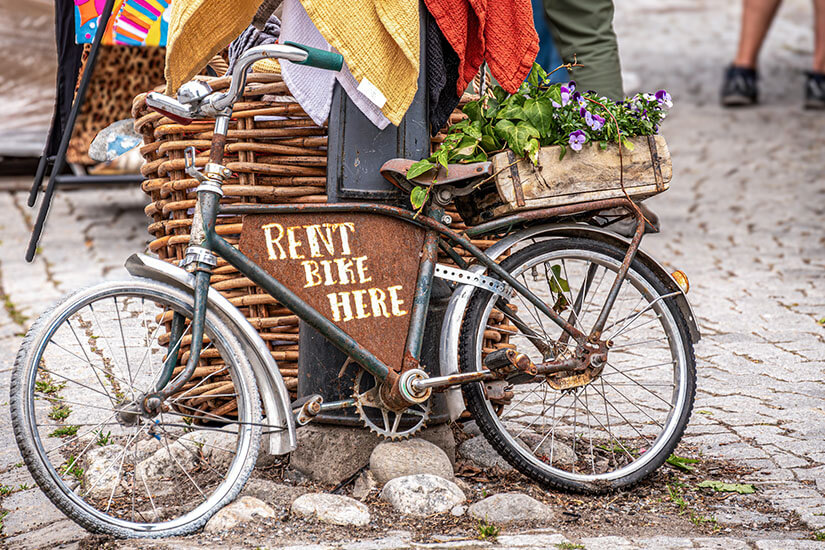 Ein altes, rostiges Fahrrad lehnt dekorativ an einem Pfosten, auf dessen Rahmen „Rent Bike Here“ steht. Hinten auf dem Gepäckträger befindet sich eine Holzkiste mit Blumen, was dem Ganzen einen charmanten Vintage-Look verleiht. Vaxholm Fahrrad