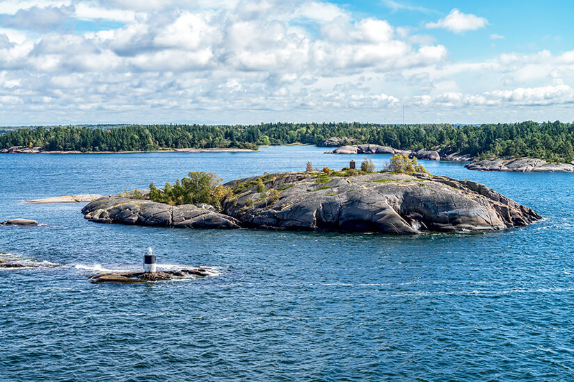 Mehrere kleine Inseln liegen im blauen Wasser, teils mit Bäumen und Sträuchern bewachsen. Im Vordergrund steht ein kleines Seezeichen auf einem einzelnen Felsen. Schweden Inselhopping