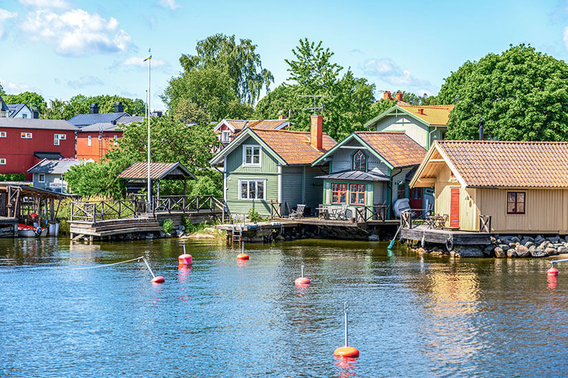 Mehrere farbige Holzhäuser stehen idyllisch am Wasser, einige davon auf Pfählen oder kleinen Stegen. Rote Bojen treiben im ruhigen Hafenbereich, der von üppigem Grün umgeben ist. Inseln Schweden Vaxholm