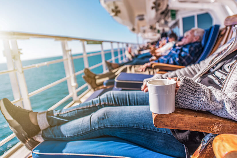 Mehrere Menschen entspannen auf Liegestühlen an Deck eines Schiffs und genießen die Sonne. Im Vordergrund hält jemand in Jeans und Strickjacke eine Tasse in der Hand. Tipps fuer die erste Kreuzfahrt Deck