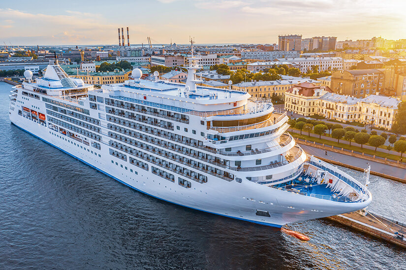 Ein großes, weißes Kreuzfahrtschiff liegt am Kai einer Stadt, die in warmem Abendlicht erstrahlt. Man erkennt mehrere Decks mit Balkonen und offenen Bereichen für Passagiere. Tipps fuer die erste Kreuzfahrt Luftaufnahme
