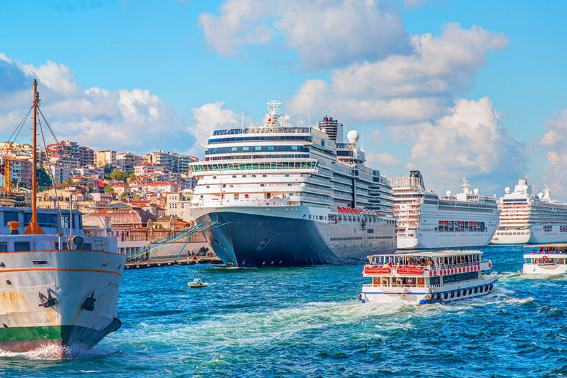Große Kreuzfahrtschiffe liegen in einem belebten Hafen vor einer farbenfrohen, hügeligen Stadtkulisse. Auf dem Wasser fahren kleinere Ausflugsboote, die weiße Wellen hinter sich lassen. Kreuzfahrtschiff Istanbul