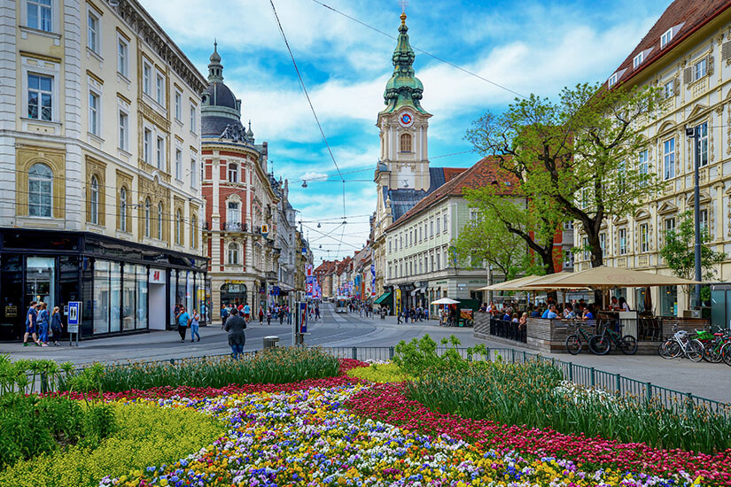Blick auf die Innenstadt von Graz. Vorne ist ein blühendes Blumenbeet zu sehen, im Hintergrund eine Straße. Oesterreich Graz