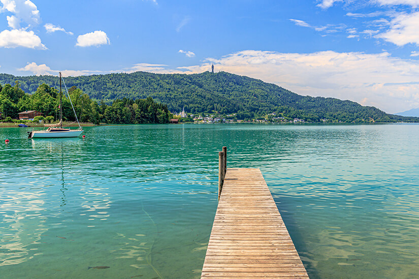 Ein Holzsteg führt über einen glaskaren See. Links ist ein Segelboot, im Hintergrund Hügel zu sehen. Kaernten Woerthersee