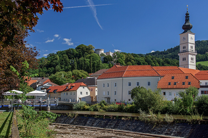 Blick über einen Fluss auf eine Stadt mit einem hohen Kirchturm. Rundherum ist grüne Vegetation zu sehen. Wolfsberg Kirche