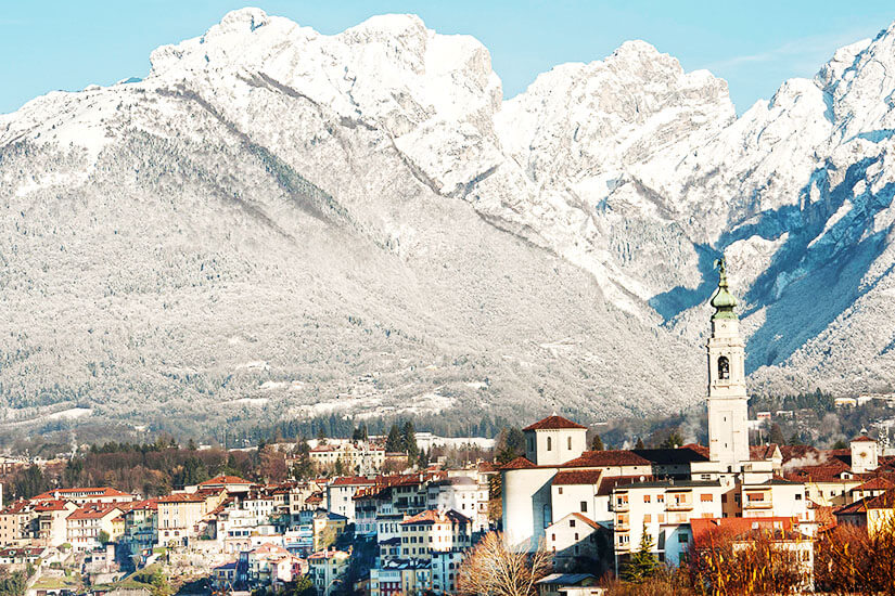 Eine Stadt vor schneebedeckten Gipfeln. Im Vordergrund ist ein Kirchturm zu sehen. Belluno Winter
